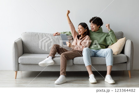 Excited young Asian couple sitting on couch with tablet pc, celebrating online win, great deal or business success at home, free space. Millennial spouses enjoying big sale in web store Excited young Asian couple sitting on couch with tablet pc, celebrating online win, great deal or business success at home, free space. Millennial spouses enjoying big sale in web store 123364482