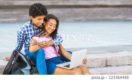 A teen couple is sitting on stone steps outside, the girl is using a laptop and guy has his arm around her. They are both smiling. The image captures a moment of intimacy and connection, copy space 123364499