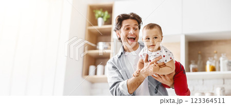 The image shows a happy father holding his baby in the kitchen. The father is smiling and looking at the camera while the baby is looking at something off-camera, copy space The image shows a happy father holding his baby in the kitchen. The father is smiling and looking at the camera while the baby is looking at something off-camera, copy space 123364511