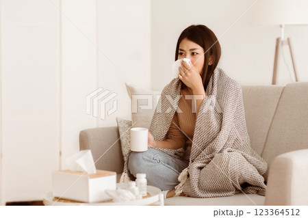 A woman sits on a couch at home, wrapped in a blanket, blowing her nose while holding a cup. She looks unwell. 123364512