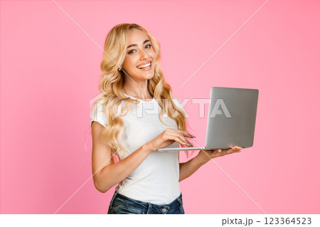 A young woman with long, blonde hair smiles while using a laptop in front of a pink background. She is wearing a white t-shirt and blue jeans. A young woman with long, blonde hair smiles while using a laptop in front of a pink background. She is wearing a white t-shirt and blue jeans. 123364523