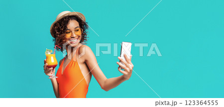 Black woman wearing a swimsuit and a straw hat smiles while taking a selfie on her smartphone. She is holding a cocktail in her other hand. The photo is taken against a bright blue background. Black woman wearing a swimsuit and a straw hat smiles while taking a selfie on her smartphone. She is holding a cocktail in her other hand. The photo is taken against a bright blue background. 123364555