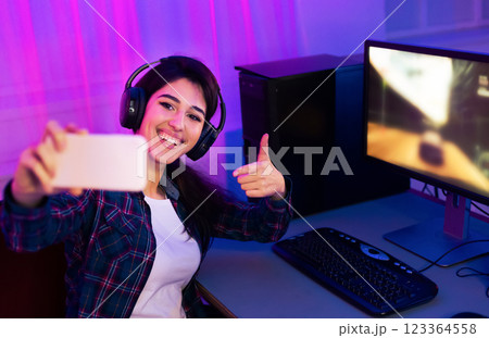 This image shows a woman sitting at a desk with a computer and keyboard. She is wearing headphones and taking a selfie with her smartphone. She is smiling and giving a thumbs-up. 123364558