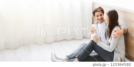 A young couple sits on the floor of their home, enjoying coffee together. The man has his arm around the woman, and they are both smiling and laughing, copy space 123364588