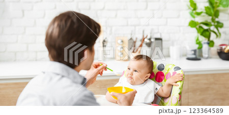 A man feeds a baby in a high chair in a kitchen. The baby is sitting in the high chair, looking at the man who is holding a spoon with food 123364589
