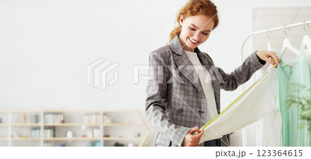 A woman in a gray blazer is examining a white blouse hanging on a rack in a clothing store. She is holding the blouse up to her body and looking at it closely. 123364615