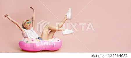 A young woman with long blonde hair, wearing a white shirt, denim shorts, a straw hat, and sunglasses, is sitting on a pink donut-shaped inflatable float in front of a pink background A young woman with long blonde hair, wearing a white shirt, denim shorts, a straw hat, and sunglasses, is sitting on a pink donut-shaped inflatable float in front of a pink background 123364621