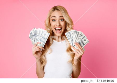 A blonde woman with a white shirt stands in front of a pink background, smiling widely and holding stacks of dollar bills in each hand. She appears to be celebrating a financial success or a win. 123364630