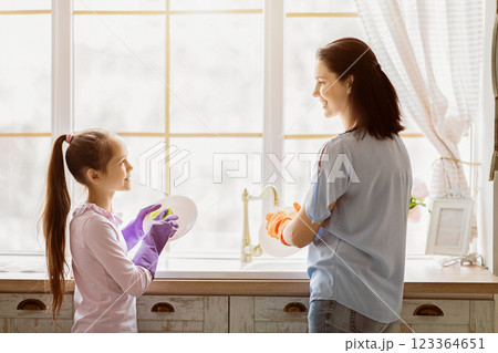 A mother and her daughter are smiling while washing dishes together in the kitchen. The two are standing near the kitchen sink and window, with the light shining through. 123364651