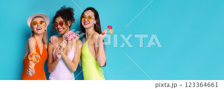 Three young women in swimsuits and sunglasses stand in front of a blue background, each holding a lollipop and smiling, copy space 123364661