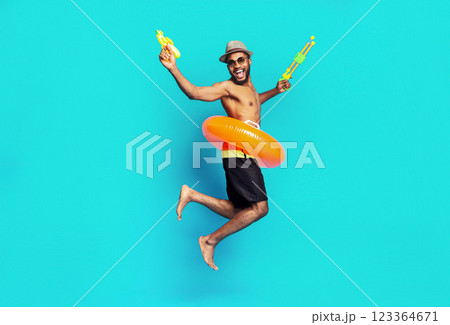 African American man is jumping in the air with a big smile on his face. He is wearing a hat, sunglasses, and swim trunks. He is holding a yellow and green water gun in each hand 123364671