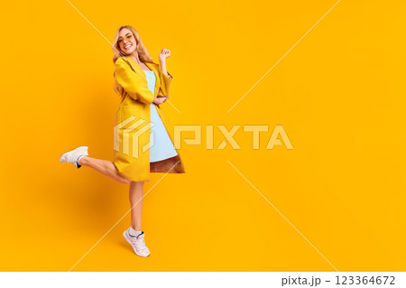 A young woman in a yellow coat and a light blue dress stands against a yellow background with one leg up and a big smile on her face. She is wearing white sneakers and has blonde hair, copy space A young woman in a yellow coat and a light blue dress stands against a yellow background with one leg up and a big smile on her face. She is wearing white sneakers and has blonde hair, copy space 123364672