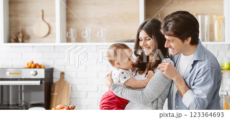 This image shows a happy family of three in their kitchen. The parents are smiling and holding their baby in their arms. The baby is looking up at the parents and appears to be happy. 123364693