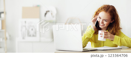 A woman sits at her desk in a modern office, talking on her cell phone, smiling, and holding a mug. A laptop sits in front of her. A woman sits at her desk in a modern office, talking on her cell phone, smiling, and holding a mug. A laptop sits in front of her. 123364706