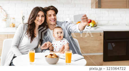A happy family of three poses for a selfie in their modern kitchen. A young mother sits at the table with her baby, while the father holds the phone and smiles at the camera, copy space A happy family of three poses for a selfie in their modern kitchen. A young mother sits at the table with her baby, while the father holds the phone and smiles at the camera, copy space 123364722