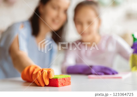 A woman wearing orange cleaning gloves holds a bright red and green sponge in her hand as she watches her daughter, who is out of focus, clean the counter. The scene is set in a kitchen. 123364725