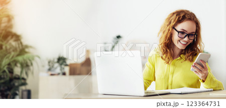A woman with red hair and glasses smiles while looking at her smartphone. She is sitting at a desk with a laptop and notepad. 123364731