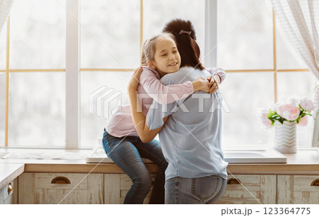 A young girl wearing jeans and a pink shirt hugs her mother from behind while standing near a window in a kitchen. They both appear to be smiling. A young girl wearing jeans and a pink shirt hugs her mother from behind while standing near a window in a kitchen. They both appear to be smiling. 123364775