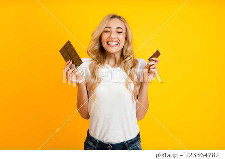 This image features a young woman with long blonde hair smiling widely while holding two chocolate bars in her hands. She is standing in front of a bright yellow background. This image features a young woman with long blonde hair smiling widely while holding two chocolate bars in her hands. She is standing in front of a bright yellow background. 123364782