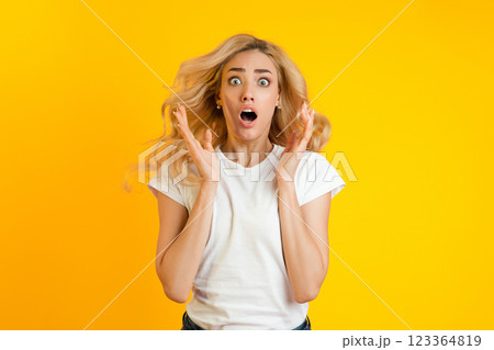 Young woman with long blonde hair wearing a white t-shirt looking shocked in front of a yellow background. Her mouth is open, her eyes are wide, and her hands are raised in a gesture of surprise. Young woman with long blonde hair wearing a white t-shirt looking shocked in front of a yellow background. Her mouth is open, her eyes are wide, and her hands are raised in a gesture of surprise. 123364819