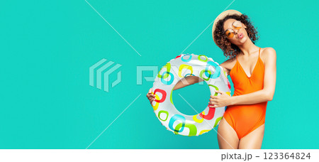 A young African American woman in an orange swimsuit stands against a turquoise background, holding a colorful inflatable ring. She is wearing a straw hat and sunglasses, copy space 123364824
