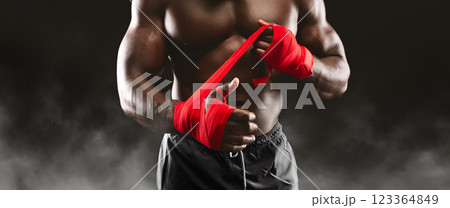 A muscular boxer prepares for a fight by wrapping his hands with red tape. A muscular boxer prepares for a fight by wrapping his hands with red tape. 123364849