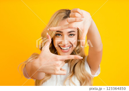 A young blonde woman with a bright smile is standing against a yellow background, making a frame with her hands. She is wearing a white t-shirt and has her hair flowing down her shoulders. A young blonde woman with a bright smile is standing against a yellow background, making a frame with her hands. She is wearing a white t-shirt and has her hair flowing down her shoulders. 123364850