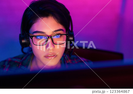 A young woman wearing glasses and headphones is intensely focused on a computer screen in a room lit by purple and blue lights. She is likely playing a video game or working on a project A young woman wearing glasses and headphones is intensely focused on a computer screen in a room lit by purple and blue lights. She is likely playing a video game or working on a project 123364867