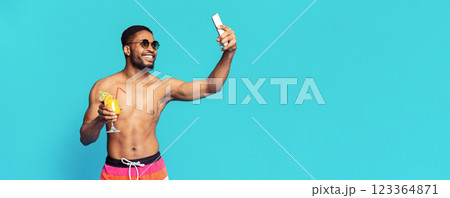 A young black man in swimwear poses for a selfie while holding a cocktail. He is wearing sunglasses and smiling. The background is a solid light blue, copy space A young black man in swimwear poses for a selfie while holding a cocktail. He is wearing sunglasses and smiling. The background is a solid light blue, copy space 123364871