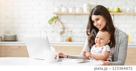 A woman sits at a table in a kitchen, working on a laptop computer. She is smiling and holding a baby in her arms, who is also smiling and looking at the camera. 123364872