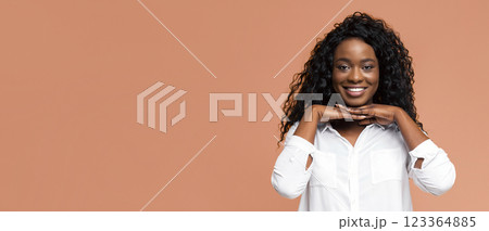 A black woman with long, curly hair is smiling at the camera while wearing a white shirt and standing in front of a peach-colored background. Her hands are cupped under her chin. 123364885