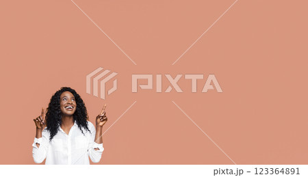 A young Black woman with curly dark hair is pointing upwards with both index fingers while smiling and looking up in a studio setting against a peach-colored backdrop. 123364891