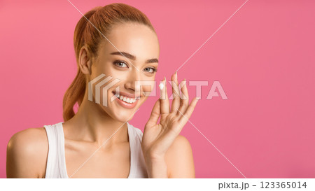 A young woman with blonde hair smiles and applies moisturizer to her cheek with her right hand. She is wearing a white tank top and standing against a plain pink background. A young woman with blonde hair smiles and applies moisturizer to her cheek with her right hand. She is wearing a white tank top and standing against a plain pink background. 123365014