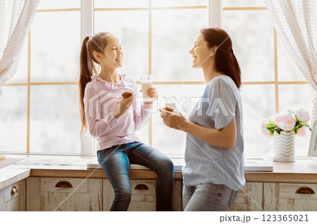A young girl and her mother are enjoying a snack break together in a kitchen, seated by a window. The girl is holding a cookie and a glass of milk, while the mother is also holding a glass of milk. 123365021