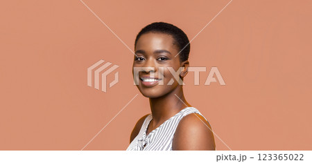 A portrait of a smiling Black woman with short hair. She is wearing a white and black striped tank top and is looking at the camera. The background is a peach color. A portrait of a smiling Black woman with short hair. She is wearing a white and black striped tank top and is looking at the camera. The background is a peach color. 123365022