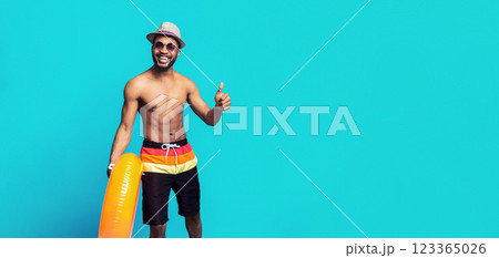 African American man in swim trunks, sunglasses, and a straw hat smiles and gives a thumbs up while holding an inflatable ring in front of a blue background. 123365026