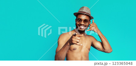 A shirtless black man with a beard and sunglasses is smiling and making a call me gesture with his right hand while pointing at the camera with his left hand against a turquoise background. A shirtless black man with a beard and sunglasses is smiling and making a call me gesture with his right hand while pointing at the camera with his left hand against a turquoise background. 123365048