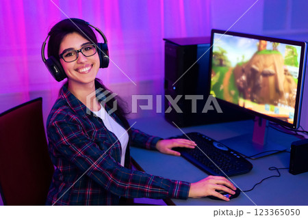 A young woman with long dark hair is smiling as she plays a computer game. She is wearing a plaid shirt and headphones. The computer monitor in the background shows an animated video game. A young woman with long dark hair is smiling as she plays a computer game. She is wearing a plaid shirt and headphones. The computer monitor in the background shows an animated video game. 123365050