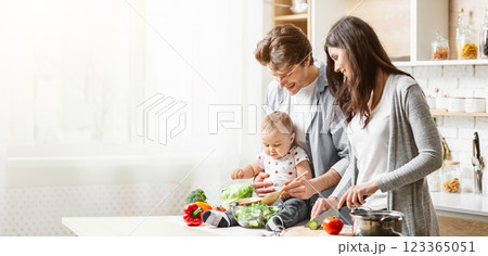 A family of three is cooking together in their kitchen. The father is holding their baby, who is sitting on the counter and reaching for a wooden spoon, copy space 123365051