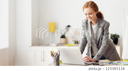 A woman in a blazer smiles as she works on a laptop in a modern office. A woman in a blazer smiles as she works on a laptop in a modern office. 123365116