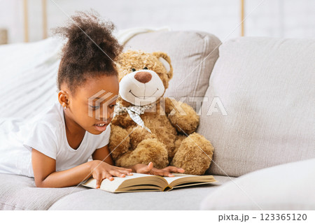 A young black girl with curly brown hair lies on a white couch, reading a book with a brown teddy bear beside her. She is wearing a white t-shirt and is smiling as she looks at the pages of the book. A young black girl with curly brown hair lies on a white couch, reading a book with a brown teddy bear beside her. She is wearing a white t-shirt and is smiling as she looks at the pages of the book. 123365120
