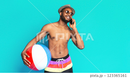 A young, smiling black man is wearing sunglasses and a hat, standing against a bright blue background. He is holding a beach ball in his left hand and talking on a phone with his right hand A young, smiling black man is wearing sunglasses and a hat, standing against a bright blue background. He is holding a beach ball in his left hand and talking on a phone with his right hand 123365161