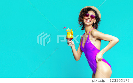 A smiling black woman is wearing a purple swimsuit and holding a tropical cocktail with a straw in one hand, while her other hand is on her hip. She is wearing sunglasses and a straw hat, copy space 123365175