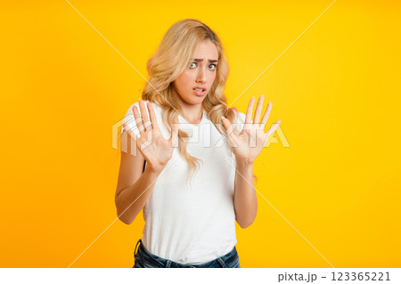 A woman with blonde hair stands in front of a yellow background, showing both hands in a stop gesture. She has a concerned expression on her face, trying to deter someone from approaching. A woman with blonde hair stands in front of a yellow background, showing both hands in a stop gesture. She has a concerned expression on her face, trying to deter someone from approaching. 123365221