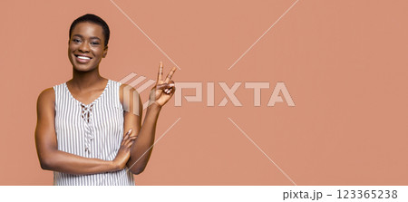 African American young woman with short hair is smiling and giving a peace sign with her right hand. She is wearing a white and black striped sleeveless top and has her left arm crossed over her torso 123365238