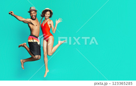 African American couple in swimwear is jumping in front of a teal background. The man is wearing black swim trunks and a straw hat, while the woman is wearing a red one-piece swimsuit and a straw hat 123365285