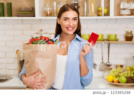A woman smiles while holding a bag of groceries and a credit card in a kitchen. 123365286