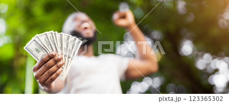 Loterry win. Happy african american man holding fan of dollars in hand outdoor, panorama 123365302