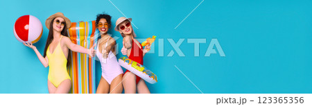 Positive young multiethnic girlfriends wearing swimsuits holding beach accessories, posing together on blue studio background, panorama with copy space 123365356