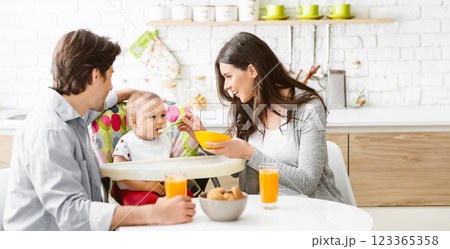 A young family enjoys a meal together in their modern kitchen. The mother is feeding their baby from a yellow bowl while the father smiles at them, copy space 123365358